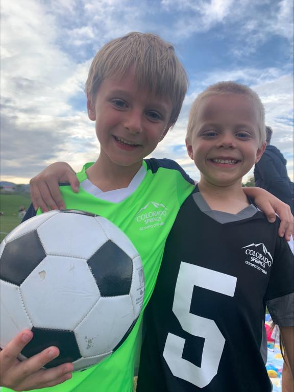 two smiling boys with their arms around each other's shoulders. They are wearing soccer jerseys and one boy is holding a soccer ball.